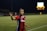 A soccer player wearing a red and blue jersey with 'GEICO' on it is on the field during a night game, gesturing with her hand. The background shows a scoreboard illuminated in the distance and another player in a yellow kit.