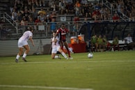 Two soccer players are focused on the ball during a match, with one player wearing a red and blue uniform and the other in a white uniform. The game takes place on a well-lit soccer field, surrounded by a crowd of spectators sitting in the stands. There are additional players and coaches seated on the sidelines, appearing attentive to the ongoing match.