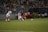 Two soccer players are focused on the ball during a match, with one player wearing a red and blue uniform and the other in a white uniform. The game takes place on a well-lit soccer field, surrounded by a crowd of spectators sitting in the stands. There are additional players and coaches seated on the sidelines, appearing attentive to the ongoing match.