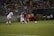 Two soccer players are focused on the ball during a match, with one player wearing a red and blue uniform and the other in a white uniform. The game takes place on a well-lit soccer field, surrounded by a crowd of spectators sitting in the stands. There are additional players and coaches seated on the sidelines, appearing attentive to the ongoing match.