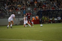 Two soccer players are focused on the ball during a match, with one player wearing a red and blue uniform and the other in a white uniform. The game takes place on a well-lit soccer field, surrounded by a crowd of spectators sitting in the stands. There are additional players and coaches seated on the sidelines, appearing attentive to the ongoing match.
