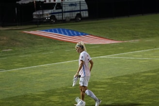 A female soccer player walks across a field carrying her cleats, with a large American flag displayed on the ground behind her. An ambulance is parked in the background near a fence.