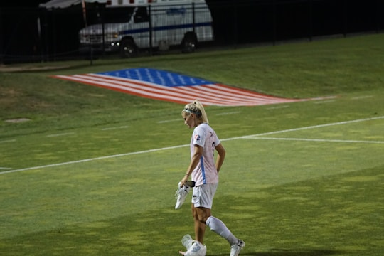 A female soccer player walks across a field carrying her cleats, with a large American flag displayed on the ground behind her. An ambulance is parked in the background near a fence.