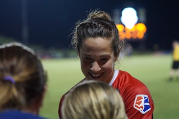 A female athlete in a red sports jersey with the NWSL logo energetically interacts with others on a sports field. The backdrop consists of a darkened night sky and a blurred scoreboard or advertisement display, with soft, colorful lights.