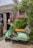A mint-green vintage scooter with a brown seat is parked on a cobblestone path in front of a rustic shop. The shop has weathered wooden siding with a bright pink window frame and a display of vibrant pink flowers. The sign above the entrance reads 'Nantucket Chic.' A mannequin with a hat and other items can be seen inside the open shop door.