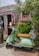 A mint-green vintage scooter with a brown seat is parked on a cobblestone path in front of a rustic shop. The shop has weathered wooden siding with a bright pink window frame and a display of vibrant pink flowers. The sign above the entrance reads 'Nantucket Chic.' A mannequin with a hat and other items can be seen inside the open shop door.