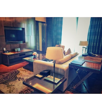 A modern, elegantly furnished living room with a large flat-screen TV mounted on the wall. There is a cozy beige sofa with decorative cushions facing a glass coffee table, flanked by two stylish side tables with lamps. A detailed patterned rug covers the wooden floor, and soft light filters through floor-length curtains.