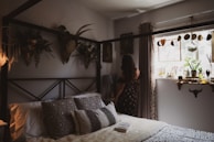 A cozy bedroom features a bed adorned with patterned pillows and a soft blanket. Wall decor includes botanical arrangements and a mounted deer head, adding a rustic touch. A woman in a floral dress stands by the window, which is decorated with plants, jars, and a dreamcatcher. Soft natural light fills the room, creating a warm atmosphere.