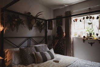 A cozy bedroom features a bed adorned with patterned pillows and a soft blanket. Wall decor includes botanical arrangements and a mounted deer head, adding a rustic touch. A woman in a floral dress stands by the window, which is decorated with plants, jars, and a dreamcatcher. Soft natural light fills the room, creating a warm atmosphere.