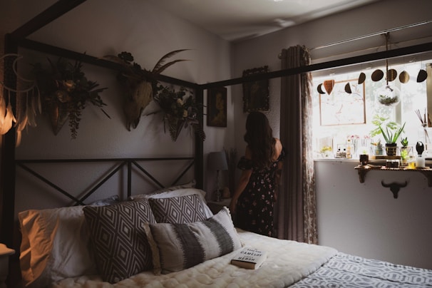 A cozy bedroom features a bed adorned with patterned pillows and a soft blanket. Wall decor includes botanical arrangements and a mounted deer head, adding a rustic touch. A woman in a floral dress stands by the window, which is decorated with plants, jars, and a dreamcatcher. Soft natural light fills the room, creating a warm atmosphere.