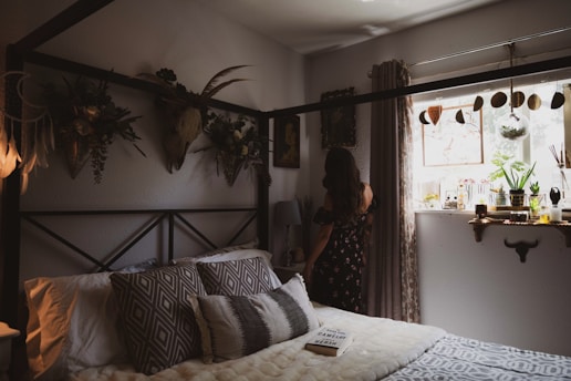 A cozy bedroom features a bed adorned with patterned pillows and a soft blanket. Wall decor includes botanical arrangements and a mounted deer head, adding a rustic touch. A woman in a floral dress stands by the window, which is decorated with plants, jars, and a dreamcatcher. Soft natural light fills the room, creating a warm atmosphere.