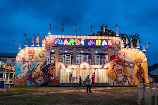 A vibrant, illuminated funhouse at a carnival with a bold Mardi Gras theme. The facade features colorful murals depicting festive and theatrical scenes, with bright lights outlining the structure. Flags add to the lively atmosphere, and a few people are gathered outside the attraction. The sky is a deep blue, suggesting evening or night time.
