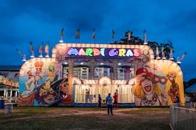 A vibrant, illuminated funhouse at a carnival with a bold Mardi Gras theme. The facade features colorful murals depicting festive and theatrical scenes, with bright lights outlining the structure. Flags add to the lively atmosphere, and a few people are gathered outside the attraction. The sky is a deep blue, suggesting evening or night time.
