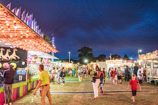 A lively carnival scene at night with various game stalls and food stands. Colorful lights illuminate the area, creating a festive atmosphere. People of different ages stroll around, some playing games and others enjoying the surroundings. A game booth with plush toys is visible in the foreground.