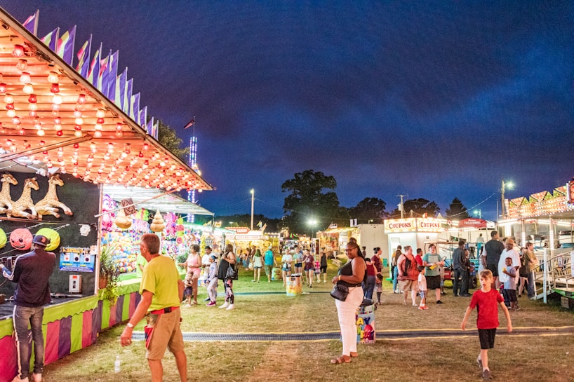 A lively carnival scene at night with various game stalls and food stands. Colorful lights illuminate the area, creating a festive atmosphere. People of different ages stroll around, some playing games and others enjoying the surroundings. A game booth with plush toys is visible in the foreground.