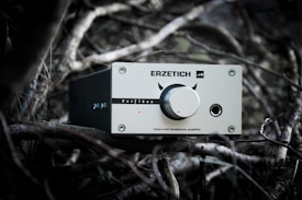 A silver headphone amplifier labeled 'ERZETICH' rests among twisted branches, featuring a central knob with decorative devil horns and various input/output ports.