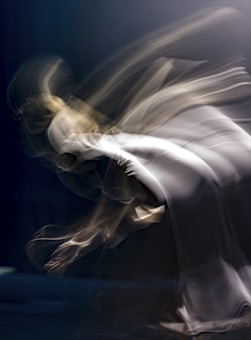 A dynamic, abstract image capturing the motion of a figure in a fluid and graceful pose. The figure is blurred, giving a sense of movement and ethereal elegance. Soft, flowing lines blend with a mixture of muted colors.