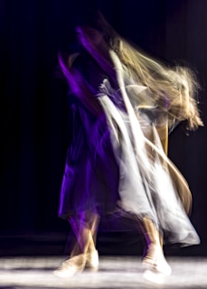 A blur of movement captures the figure of a dancer in motion, with swirling fabrics and dynamic lighting. The scene is awash with deep purple and soft beige tones, suggesting a graceful and energetic performance.