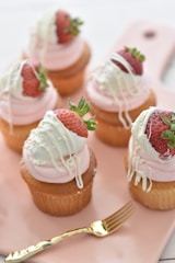 Five cupcakes are arranged on a light pink tray. Each cupcake is adorned with a swirl of pink frosting, topped with a fresh strawberry dipped in white chocolate and stylishly drizzled with additional white chocolate. A small golden fork accompanies the setup, enhancing the elegance and gourmet appeal.
