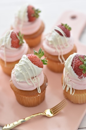 Five cupcakes are arranged on a light pink tray. Each cupcake is adorned with a swirl of pink frosting, topped with a fresh strawberry dipped in white chocolate and stylishly drizzled with additional white chocolate. A small golden fork accompanies the setup, enhancing the elegance and gourmet appeal.