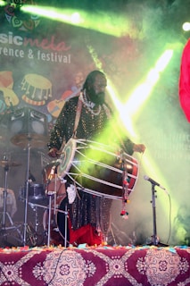 A musician is performing on stage, playing a large traditional drum. The stage is decorated with colorful lights and a backdrop with patterns and text. The musician is dressed in vibrant, traditional attire, and there are additional musical instruments, such as a drum set, visible in the background.