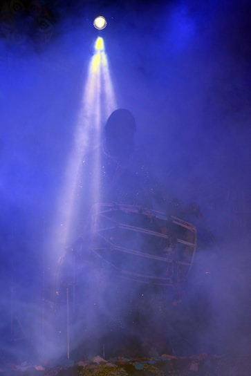 A mysterious figure is immersed in a foggy atmosphere under a spotlight. The silhouette of the person can be seen holding a large drum, and the light creates an ethereal glow around them.