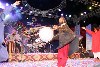 A lively musical performance is taking place on stage, featuring a group of musicians. A man in traditional attire is energetically playing a large drum, while another person is visible in the background playing a smaller percussion instrument. There is a vibrant atmosphere with colorful decorations and confetti scattered on the stage floor. Bright stage lights illuminate the scene, adding to the festive mood.