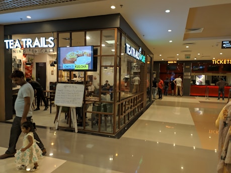 A shopping mall hallway with a cafe on the left named 'Tea Trails Dining Tea Cafe' with a digital sign displaying 'Choco Kulcha'. People are visible inside the cafe and some sitting by the glass walls. A man and a young girl are walking in the foreground. On the right side, there is a ticket booth with people queueing.