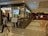 A shopping mall hallway with a cafe on the left named 'Tea Trails Dining Tea Cafe' with a digital sign displaying 'Choco Kulcha'. People are visible inside the cafe and some sitting by the glass walls. A man and a young girl are walking in the foreground. On the right side, there is a ticket booth with people queueing.