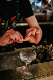 A bartender prepares a cocktail, squeezing a lemon peel over a chilled coupe glass filled with clear liquid. The bar counter is dotted with a perforated surface, and the background reveals blurred bottles and bar equipment. The bartender wears a dark shirt with a colorful apron.