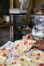 A freshly baked pizza with slices of melted cheese, cherry tomatoes, and herbs is being served. A hand is holding one slice, ready to take it from the tray lined with branded paper. The background features a glass display counter and blurred figures, suggesting a bustling café or pizzeria setting.