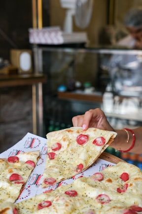 A freshly baked pizza with slices of melted cheese, cherry tomatoes, and herbs is being served. A hand is holding one slice, ready to take it from the tray lined with branded paper. The background features a glass display counter and blurred figures, suggesting a bustling café or pizzeria setting.