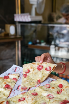A freshly baked pizza with slices of melted cheese, cherry tomatoes, and herbs is being served. A hand is holding one slice, ready to take it from the tray lined with branded paper. The background features a glass display counter and blurred figures, suggesting a bustling caf&eacute; or pizzeria setting.