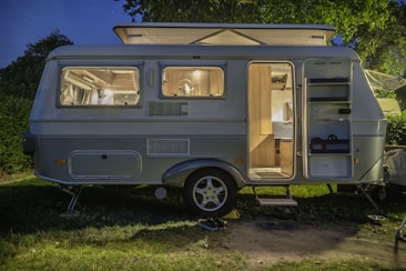 A cozy caravan is parked on grass with its interior warmly lit. The open door reveals a neatly arranged space, including a bed and kitchen area. The caravan is set under a tree, surrounded by greenery.