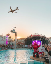 A vibrant outdoor party at a poolside venue with a large crowd gathered around a stage. A plane is flying overhead in the clear sky, and a creative water feature in the form of a giant floating tap is pouring water into the pool. Bright lights and a colorful digital screen on the stage add to the energetic atmosphere.