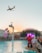 A vibrant outdoor party at a poolside venue with a large crowd gathered around a stage. A plane is flying overhead in the clear sky, and a creative water feature in the form of a giant floating tap is pouring water into the pool. Bright lights and a colorful digital screen on the stage add to the energetic atmosphere.