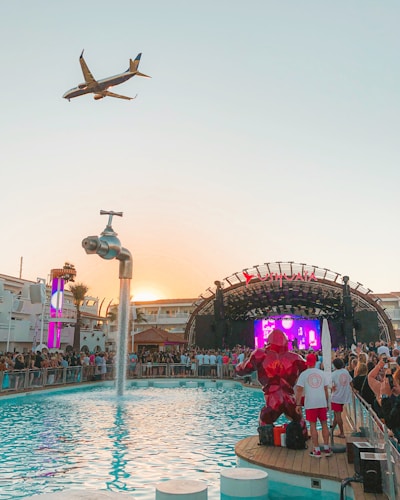 A vibrant outdoor party at a poolside venue with a large crowd gathered around a stage. A plane is flying overhead in the clear sky, and a creative water feature in the form of a giant floating tap is pouring water into the pool. Bright lights and a colorful digital screen on the stage add to the energetic atmosphere.