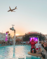 A vibrant outdoor party at a poolside venue with a large crowd gathered around a stage. A plane is flying overhead in the clear sky, and a creative water feature in the form of a giant floating tap is pouring water into the pool. Bright lights and a colorful digital screen on the stage add to the energetic atmosphere.