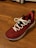 A single red athletic shoe with white laces is placed on a wooden floor. The shoe features a textured upper with a decorative emblem on the side and black eyelets.