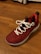 A single red athletic shoe with white laces is placed on a wooden floor. The shoe features a textured upper with a decorative emblem on the side and black eyelets.