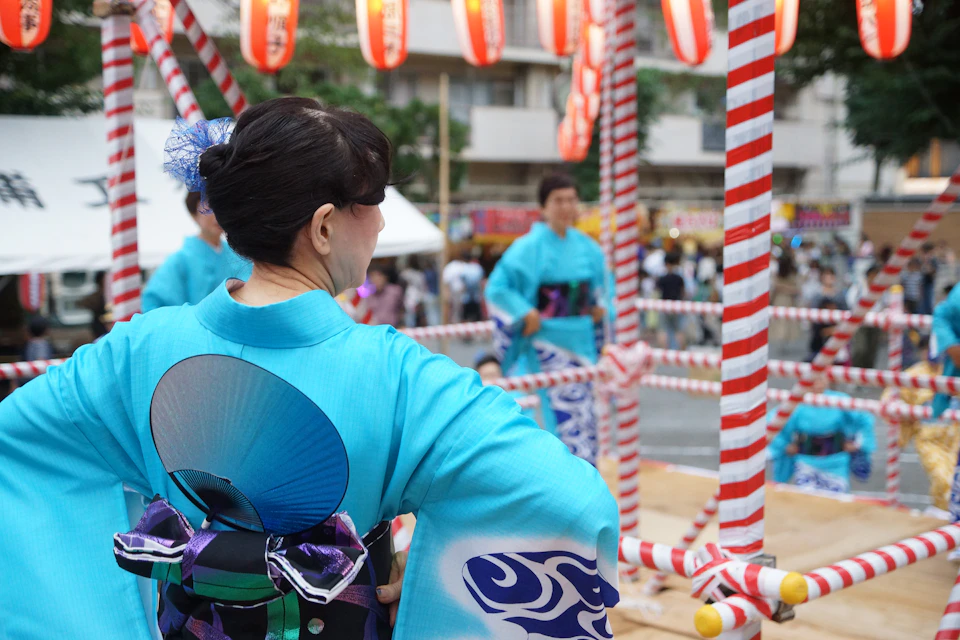 A woman in a pale blue yukata with a fan tucked into her obi at a bon odori, with striped poles and paper lanterns around the dance floor