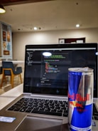 A laptop displaying code on its screen, set on a table in a modern, well-lit cafe. An open can of Red Bull energy drink is placed prominently next to the laptop. In the background, there are chairs and decorative wall posters.