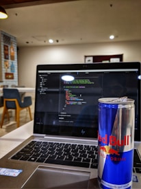 A laptop displaying code on its screen, set on a table in a modern, well-lit cafe. An open can of Red Bull energy drink is placed prominently next to the laptop. In the background, there are chairs and decorative wall posters.