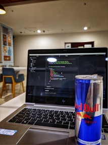 A laptop displaying code on its screen, set on a table in a modern, well-lit cafe. An open can of Red Bull energy drink is placed prominently next to the laptop. In the background, there are chairs and decorative wall posters.