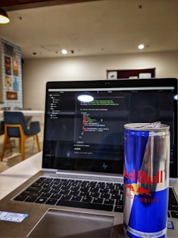 A laptop displaying code on its screen, set on a table in a modern, well-lit cafe. An open can of Red Bull energy drink is placed prominently next to the laptop. In the background, there are chairs and decorative wall posters.