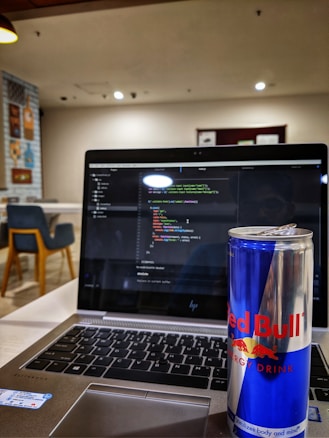 A laptop displaying code on its screen, set on a table in a modern, well-lit cafe. An open can of Red Bull energy drink is placed prominently next to the laptop. In the background, there are chairs and decorative wall posters.