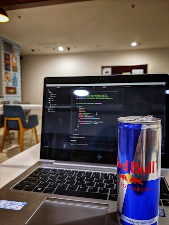 A laptop displaying code on its screen, set on a table in a modern, well-lit cafe. An open can of Red Bull energy drink is placed prominently next to the laptop. In the background, there are chairs and decorative wall posters.