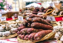 A market stall features a variety of sausages displayed in wicker baskets, with small signs indicating different types. The background includes more baskets and blurred flowers. The red and white checkered tablecloth adds a rustic feel.