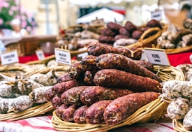 A market stall features a variety of sausages displayed in wicker baskets, with small signs indicating different types. The background includes more baskets and blurred flowers. The red and white checkered tablecloth adds a rustic feel.