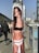A poster of a young woman wearing a black top and white underwear with the text 'HEROIN KIDS' is wrapped around a pole on a city street. In the background, there are blurred images of people walking and urban infrastructure, including buildings and vehicles. Graffiti is visible on an electrical box nearby.